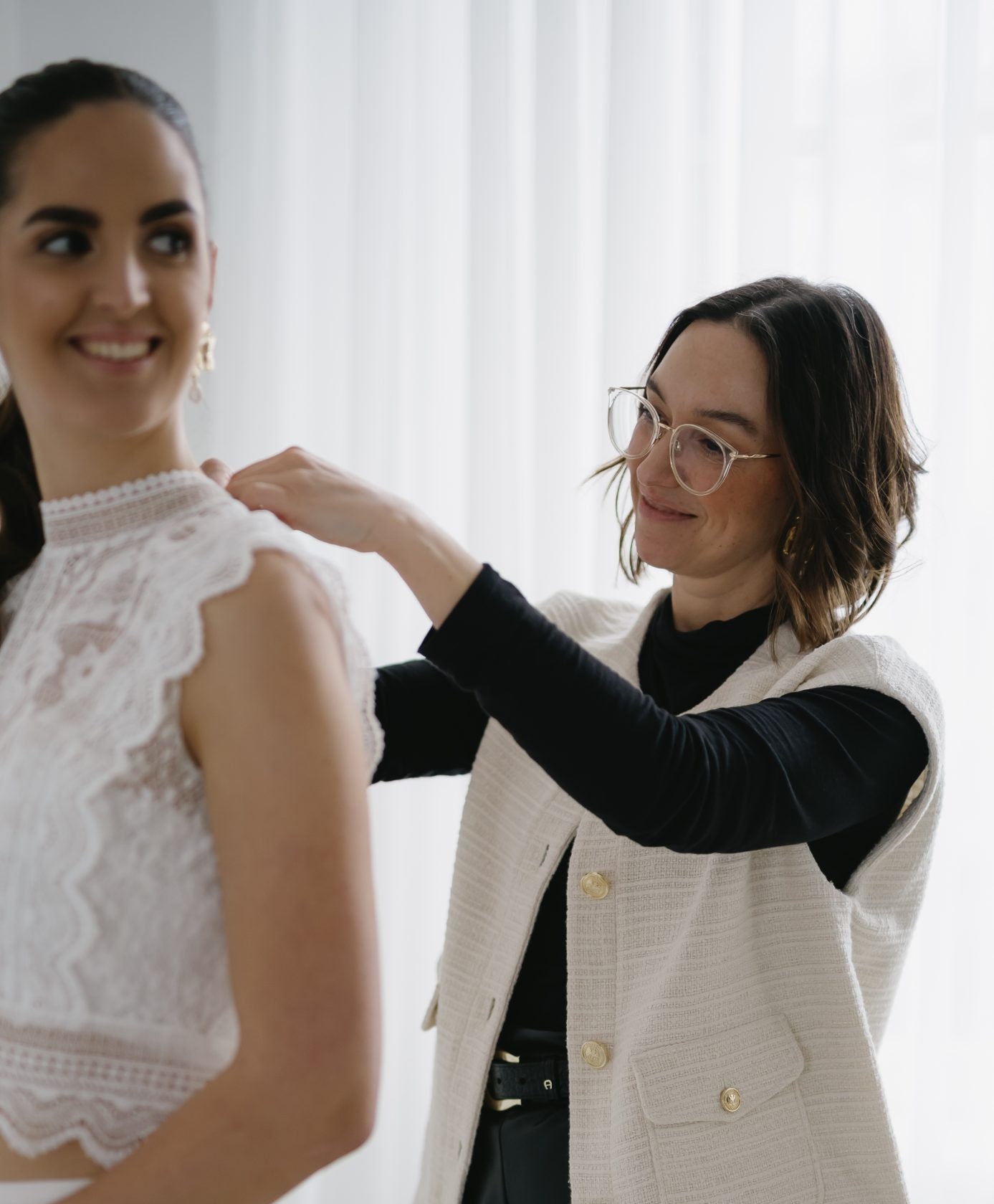 Eine Frau hilft einer anderen Frau, ihr Spitzenkleid in einem hellen Raum mit weißen Vorhängen zurechtzurücken. Eine elegante Szene, die sich perfekt für ein Archiv oder ein Elementor-Portfolio eignet.