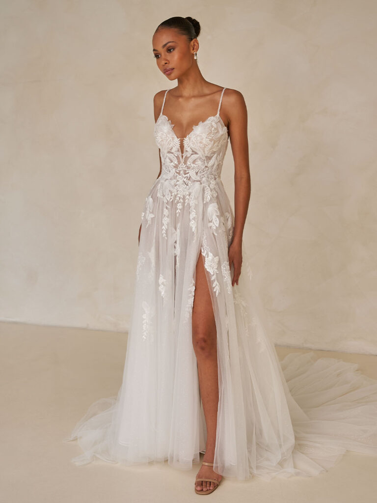 Frau in einem weißen Hochzeitskleid aus floraler Spitze mit dünnen Trägern und hohem Schlitz, stehend in einem Haus.