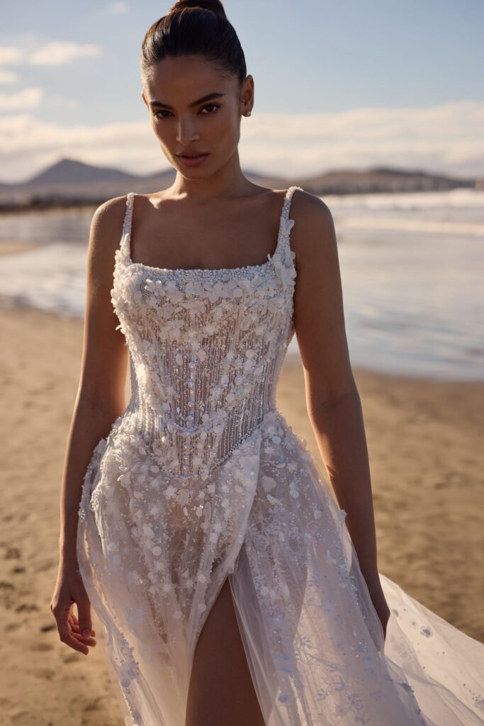 Eine Frau in einem blumengeschmückten, weißen Brautkleid steht an einem Sandstrand mit Wasser und Bergen im Hintergrund und zeigt elegante, von Fulda inspirierte Brautmode.