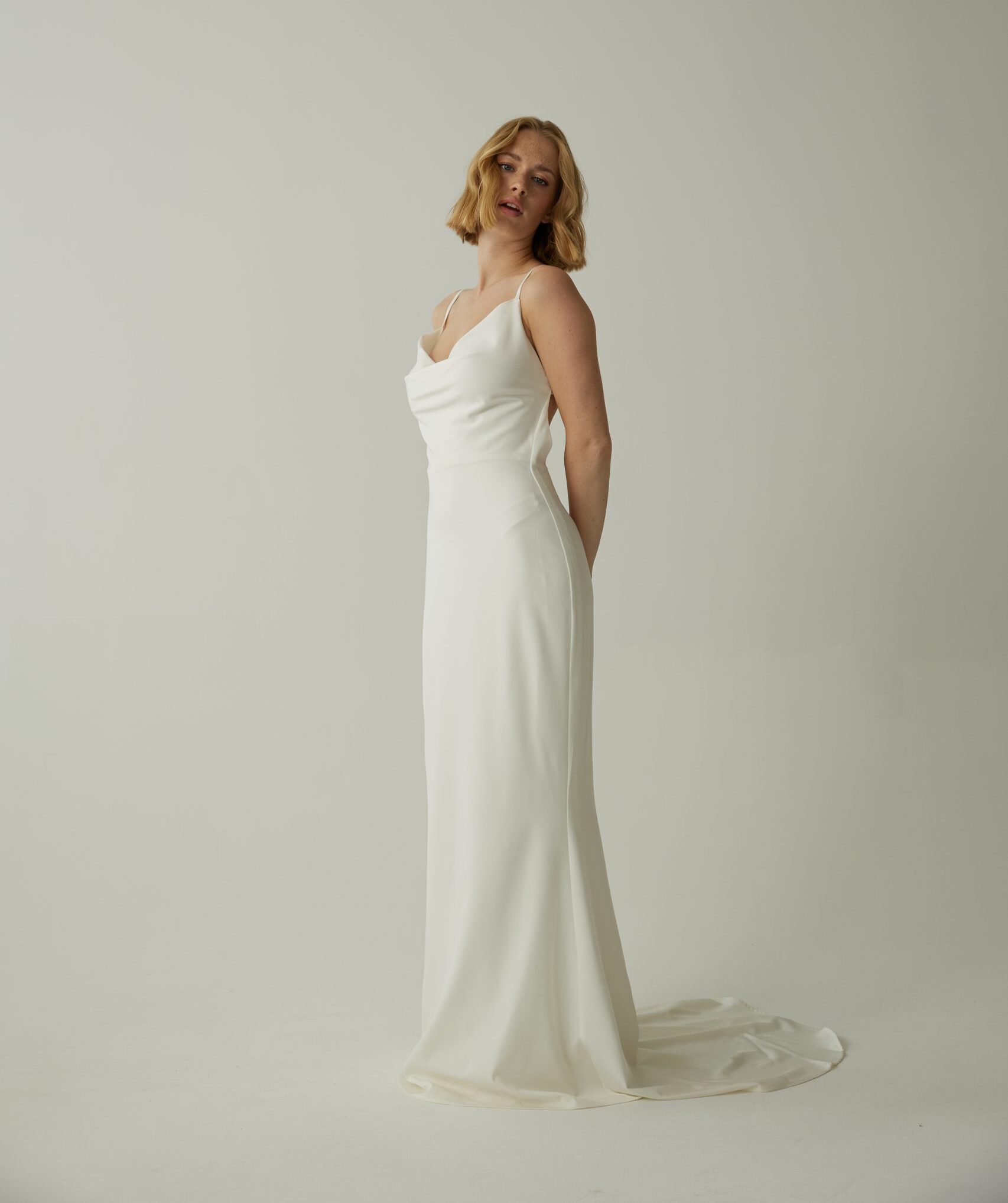 Frau in einem ärmellosen weißen Brautkleid mit Schleppe, stehend vor einem schlichten hellen Hintergrund.