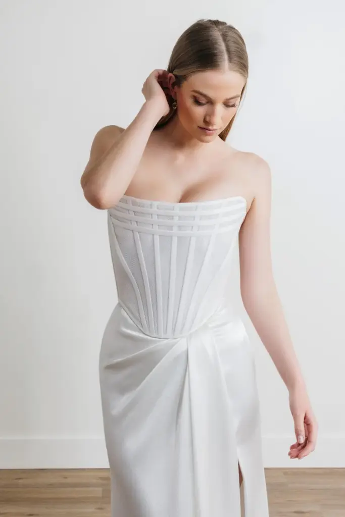 Nayeli steht drinnen in einem trägerlosen weißen Abendkleid mit strukturiertem Oberteil und drapiertem Satinrock und strahlt zeitlose Eleganz aus, während dezente 68716B-Akzente dem Outfit einen modernen Touch verleihen.