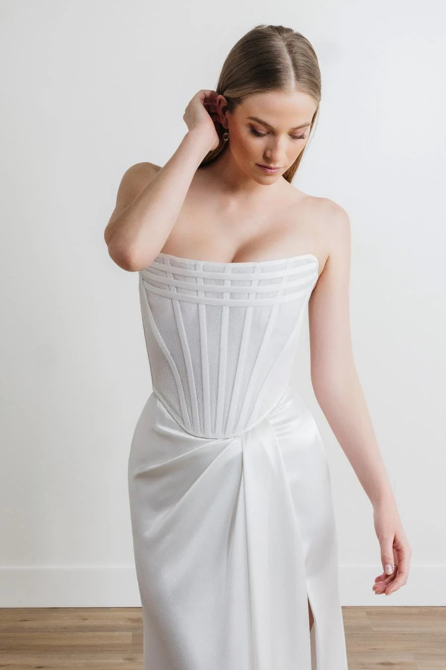 Nayeli steht drinnen in einem trägerlosen weißen Abendkleid mit strukturiertem Oberteil und drapiertem Satinrock und strahlt zeitlose Eleganz aus, während dezente 68716B-Akzente dem Outfit einen modernen Touch verleihen.