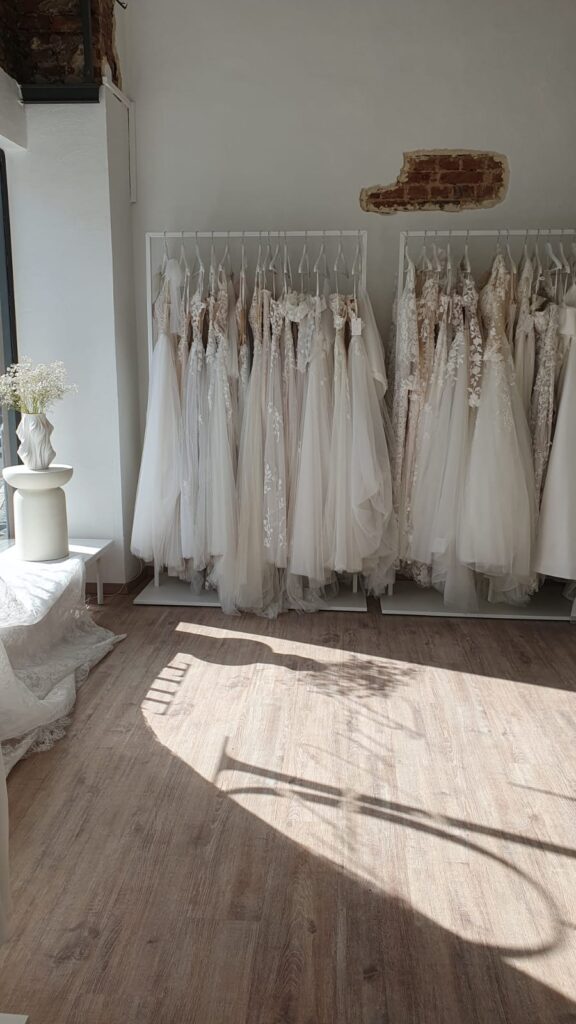 Brautkleider, die in einer sonnendurchfluteten Brautmodenboutique mit Holzböden und weißen Wänden an Kleiderständern hängen.