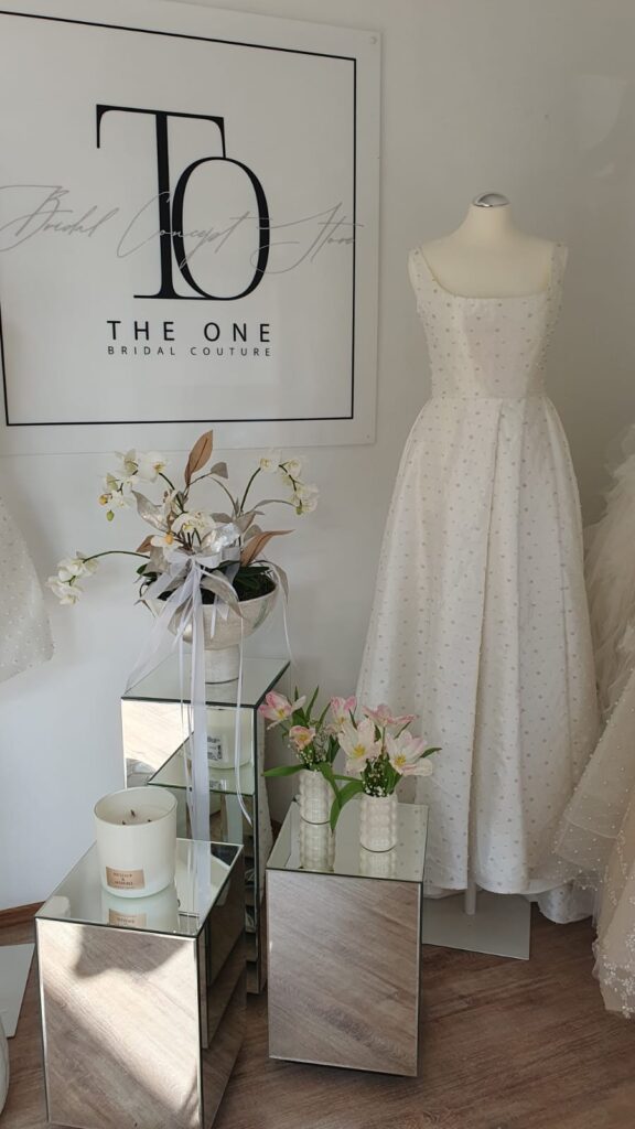 Ein weißes Brautkleid auf einer Schaufensterpuppe neben Blumen, Kerzen und einem Schild mit der Aufschrift „THE ONE BRIDAL COUTURE“.