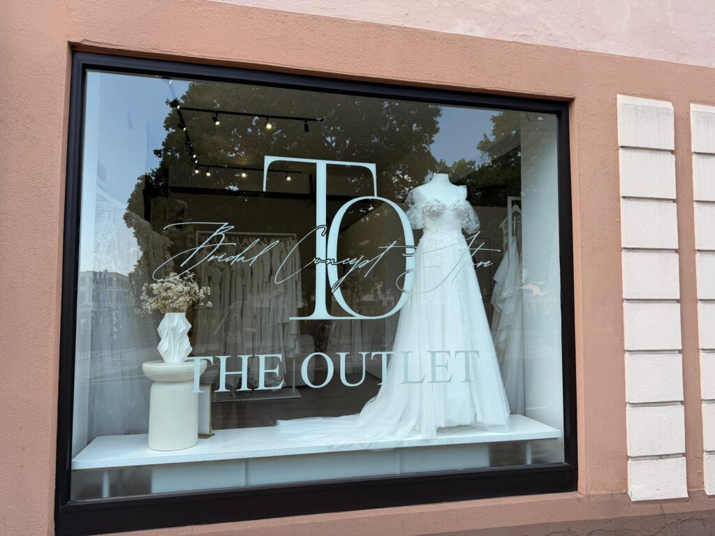 Schaufensterauslage mit einem weißen Brautkleid und einer Vase mit Blumen im Brautmodengeschäft „The Outlet“.