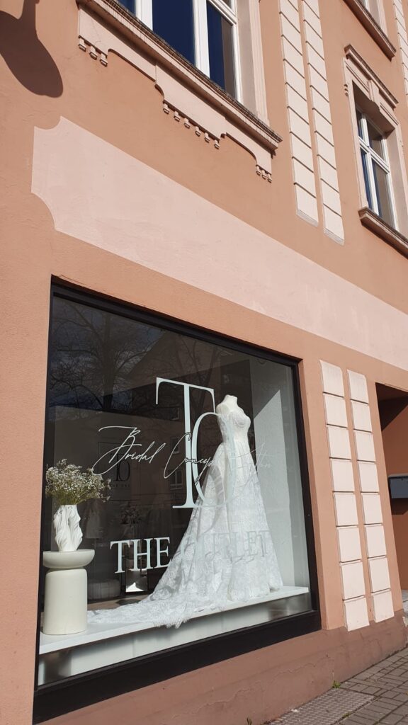 Im Schaufenster eines Brautmodengeschäfts ist ein weißes Brautkleid auf einer Schaufensterpuppe neben einer Vase mit weißen Blumen ausgestellt.