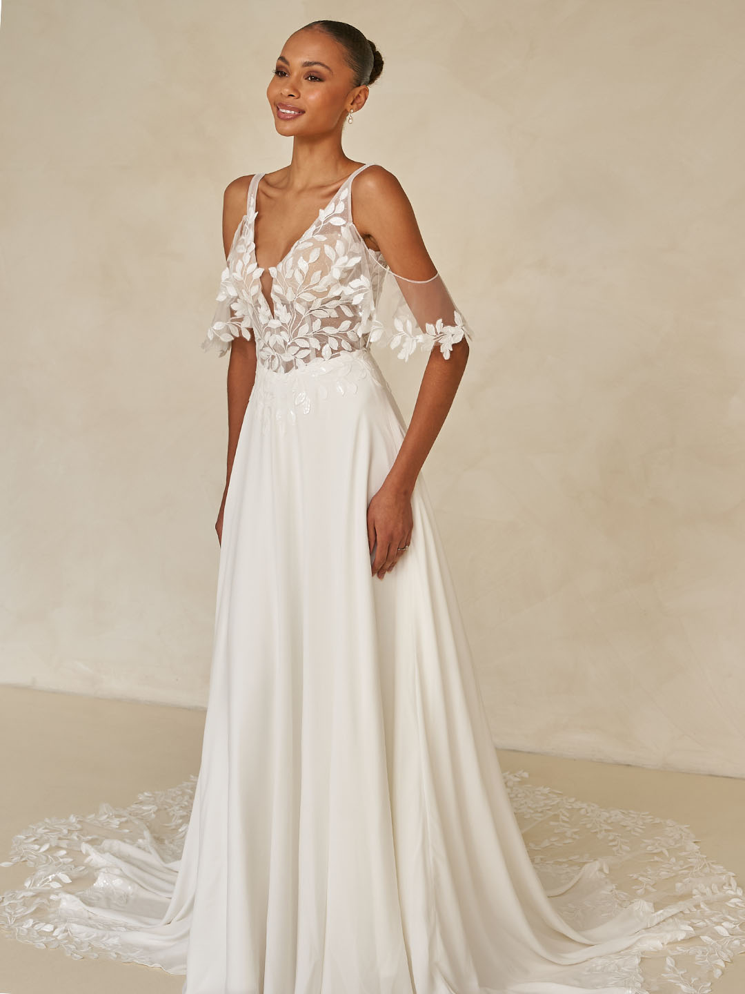 Eine Frau in einem eleganten weißen Brautkleid mit einem Oberteil aus Blumenspitze, transparenten Ärmeln und einem fließenden Rock.