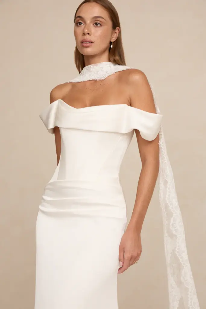 Eine Frau in einem weißen, schulterfreien Abendkleid, über eine Schulter drapiert ein Spitzenschal, posiert vor einem beigen Hintergrund.