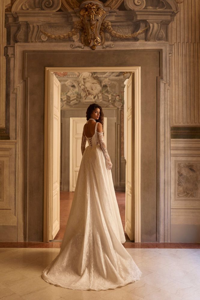 Eine Braut in einem langen weißen Kleid steht in einem prunkvollen Flur und hat den Rücken zu den kunstvollen Deckenverzierungen gewandt.