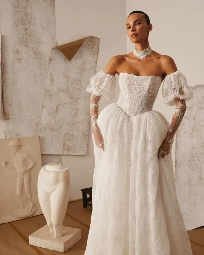 Eine Frau in einem weißen, schulterfreien Brautkleid mit transparenten Ärmeln, die neben abstrakten Skulpturen und Kunstwerken steht.